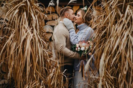Newlyweds stand near dry grass on background of firewood stack and embrace.