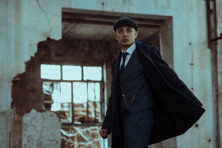 A man posing in the image of an English retro gangster of the 1920s dressed in a coat, suit and flat cap in Peaky blinders style.の写真素材