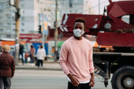 A young African man stands with a medical protective mask on his face at city street. Concept of preventive measures and protection for coronavirus pandemic.の写真素材