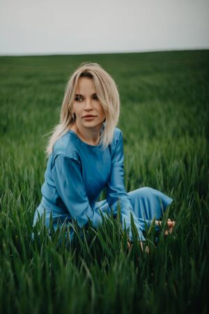 A young woman in blue dress sits among the green field of wheat.