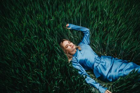 Young woman in a blue dress lies, relaxes and rests among green wheat. Top view. Copy space.