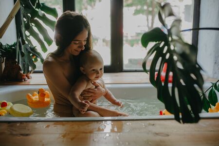 Mother with her baby bathe in bathtub on background of houseplants and floating toys.の写真素材