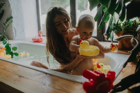 Mother with her baby bathe in bathtub and play with floating toys on houseplants background.の写真素材