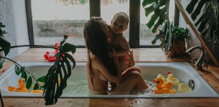 Mother hugs and kisses her baby while bathing in bathtub with floating toys.の写真素材