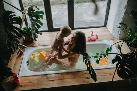 Mother bathes in bathtub and plays with her baby on background of floating toys and houseplants.の写真素材
