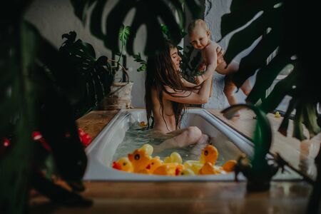 Mother bathes in bathtub and plays with her baby on background of floating toys and houseplants.の写真素材