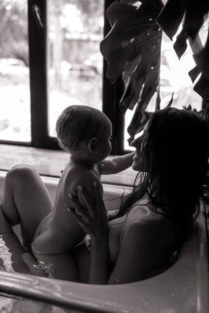 Mother bathes in bathtub and plays with her baby on background of window. Black and white image.の写真素材