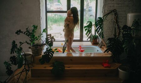 Mother stands with baby in her arms in bathtub full water and floating toys on window background.の写真素材