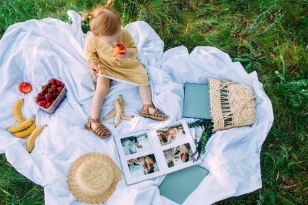 Child girl sits, looks photo album and eats fruits during picnic in the park.の写真素材