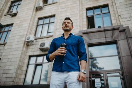 Young man in jeans and shirt stands with cup of coffee in his hands against background of modern building. Bottom view.の写真素材