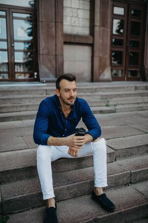 Young man sits and dreams on stairs with cup of coffee in his hands against background of modern...