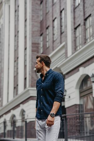 Young man in jeans and shirt looks around on street against background of modern building.の写真素材