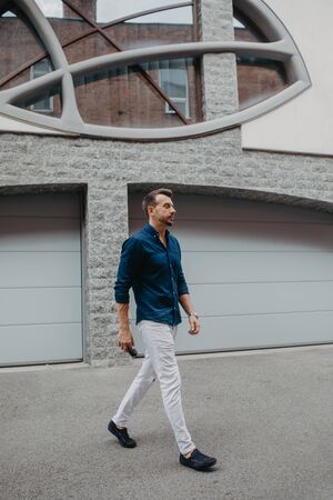 Young man in jeans and shirt goes at street with sunglasses in his hands against background of building.の写真素材