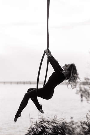 Woman aerialist performs acrobatic elements in hanging aerial hoop against background of river. Black and white image.の写真素材