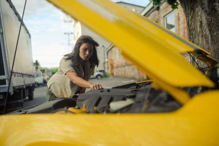 Young woman stands near yellow car with opened bonnet and repairs it against background of building.