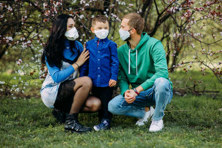 Happy family with little son are in a medical protective respirators on their faces against background of blooming spring garden during the walk. Concept of preventive measures and protection for coronavirus pandemic.の写真素材