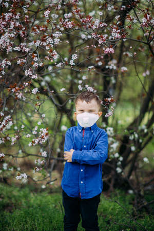 Happy child boy stands with a medical protective respirator on his face against background of blooming spring garden during the walk. Concept of preventive measures and protection for coronavirus pandemic.の写真素材