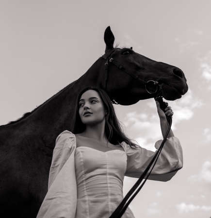 Young woman stands and holds horse by bridle against sky background. Black and white image.の写真素材