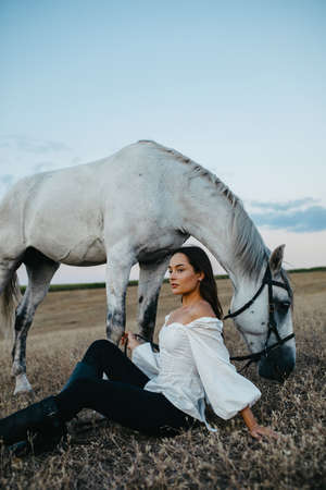 Young pensive woman sits near gray horse in pasture.の写真素材