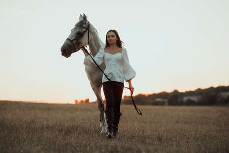 Young woman leads a white horse by the bridle through the pasture at sunset.