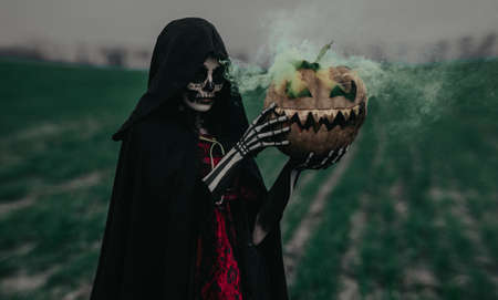 Young woman stands among field in Halloween costume of death with painted skeleton on her body and holds jack-o-lantern in her hands with yellow smoke from it. Halloween concept. Sugar skull makeup.の写真素材