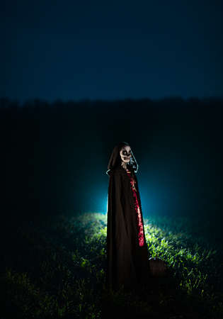 Young woman stands among field in Halloween costume of death with painted skeleton on her body against dark sky. Halloween concept. Sugar skull makeup. Backlit.の写真素材