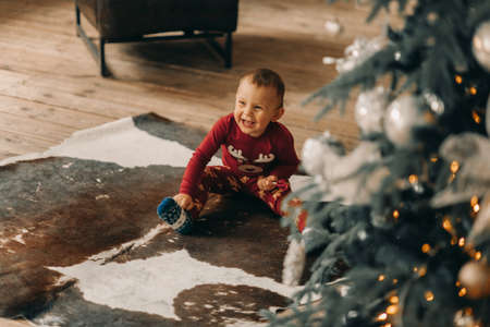 Boy sits on carpet in suit with Christmas ornament and has fun near Christmas tree.の写真素材