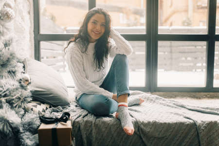 Happy young woman sits on windowsill near Christmas tree and box with gift.