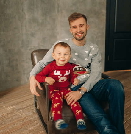 Father with his son sit in armchair in sweaters with Christmas ornament.