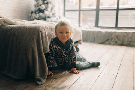 Happy child boy sits on the floor near bed against Christmas tree and window.