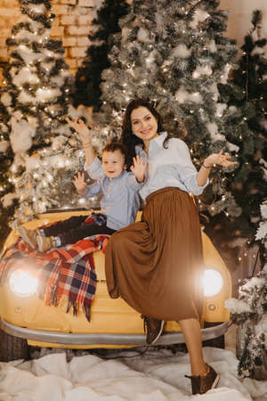 Mother and her son sit on a retro car hood and have a fun against background of Christmas tree and decorations.の写真素材