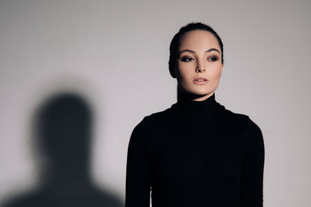Portrait of young woman in black turtleneck next her shadow against of gray background.の写真素材