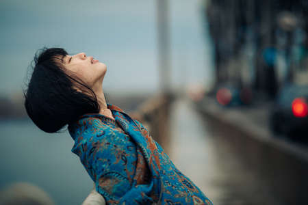 Portrait of young Japanese relaxed woman wrapped in scarf on the bridge against background of river and cars.の写真素材