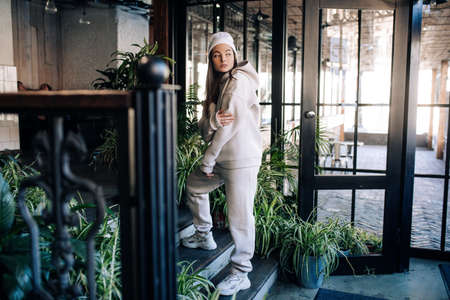 Young woman in sweatsuit stands on the stairs of the porch in front of the entrance to the house.