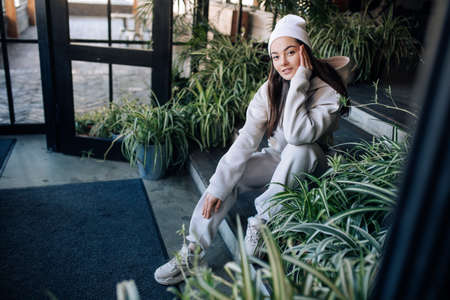 Young woman in sweatsuit sits on the stairs of the porch in front of the entrance to the house.