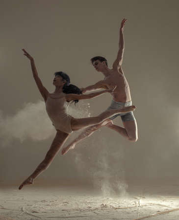Two ballet dancers perform dance and jump in splits and twine poses above the floor covered flour...