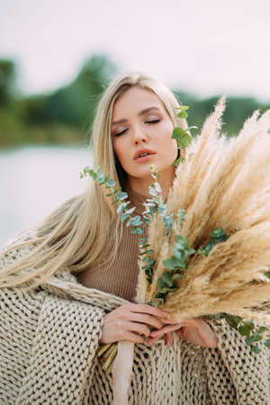 Young blonde woman poses on beach near lake with bouquet of dry flowers in her hands. Portrait.の写真素材