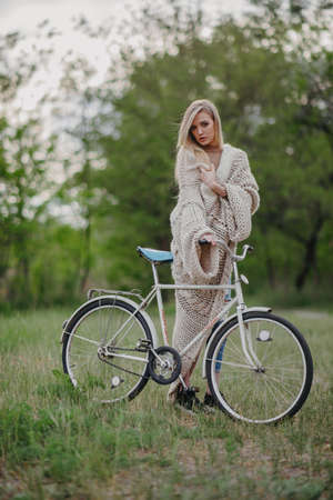 Young blonde woman stands among meadow and holds bicycle in her hands.