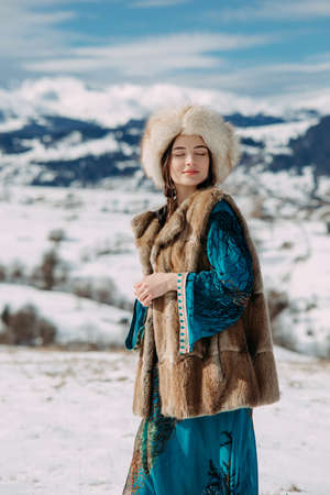 Young woman walks dressed in fur hat, vest and dress against beautiful landscape of snowbound Carpathians mountains and sky.の写真素材