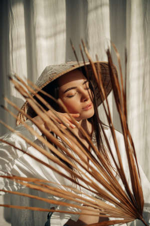 Portrait of young sunlit woman in white shirt and vietnamese hat with closed eyes.の写真素材