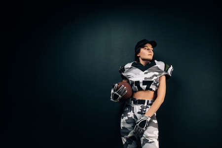 Beautiful sexy athletic woman poses on black background in American football uniform and cap with ball in her hands. Copy spaceの写真素材