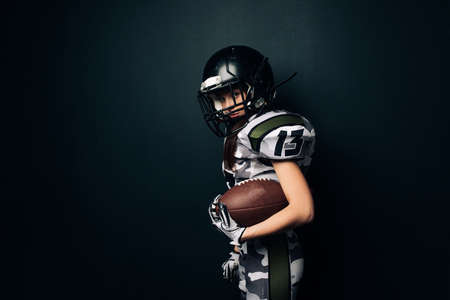 Beautiful sexy athletic woman poses on black background in American football uniform and helmet with ball in her hand. Side view.の写真素材