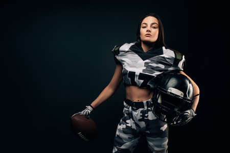 Beautiful sexy athletic woman poses on black background in American football uniform with helmet and ball in her hands.の写真素材