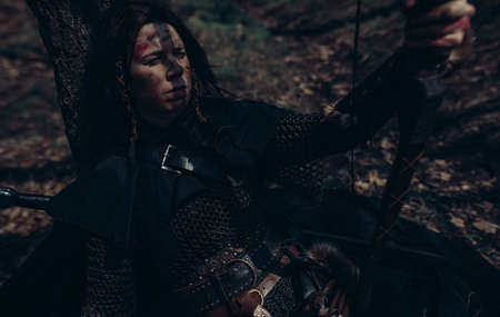 Woman in chain mail in image of medieval warrior sits and rests near tree among forest. Closeup.の写真素材