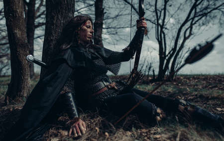 Woman in chain mail in image of medieval warrior sits and rests near tree among forest.の写真素材