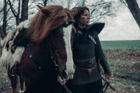 Woman in chain mail in image of medieval warrior stands near horse and holds it by the reins among forest.の写真素材