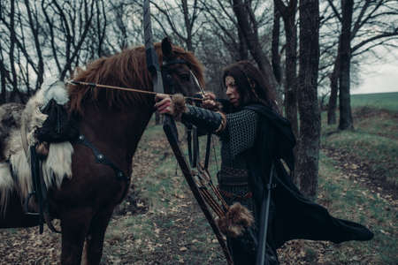 Woman in image of medieval warrior archer shoots a bow and aims at target near her horse among...