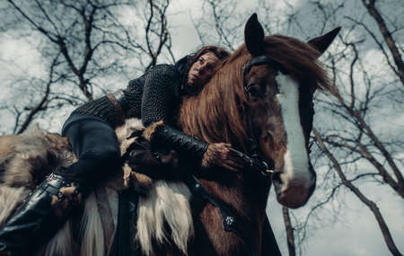 Woman in chain mail in image of medieval warrior sits horseback and embraces it among forest.の写真素材