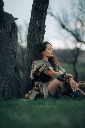 Woman in image of ancient warrior amazon sits and rests under tree on grass in forest.