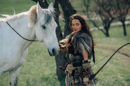 Woman stands in fur clothes in image of ancient warrior amazon with bow in her hands in forest near...